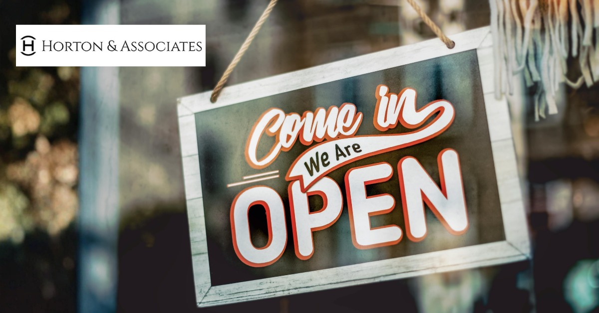 A vintage wooden sign reading Come in We Are Open hanging in a storefront window next to a Horton and Associates logo.